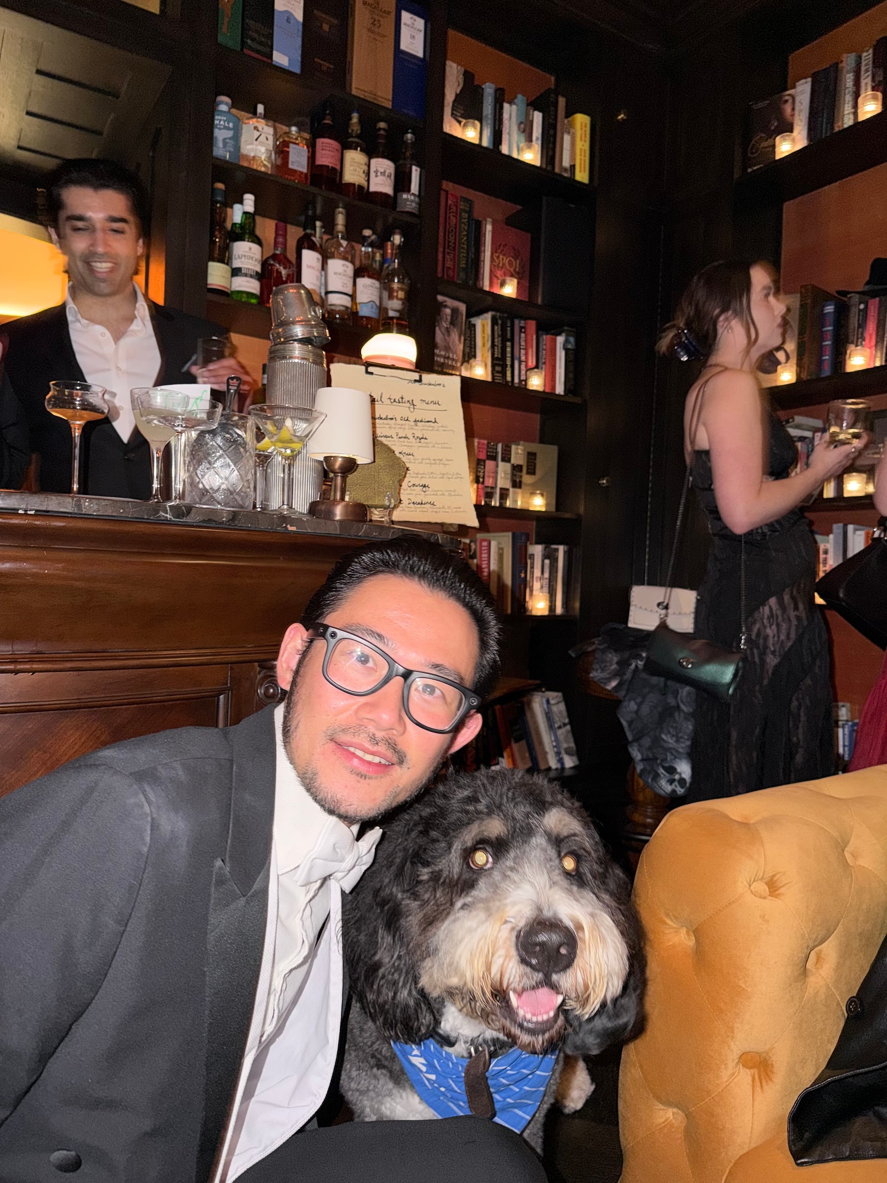 Jay crouching next to a fluffy black, gray and white doodle wearing a blue bandana in a bookshelf-lined bar