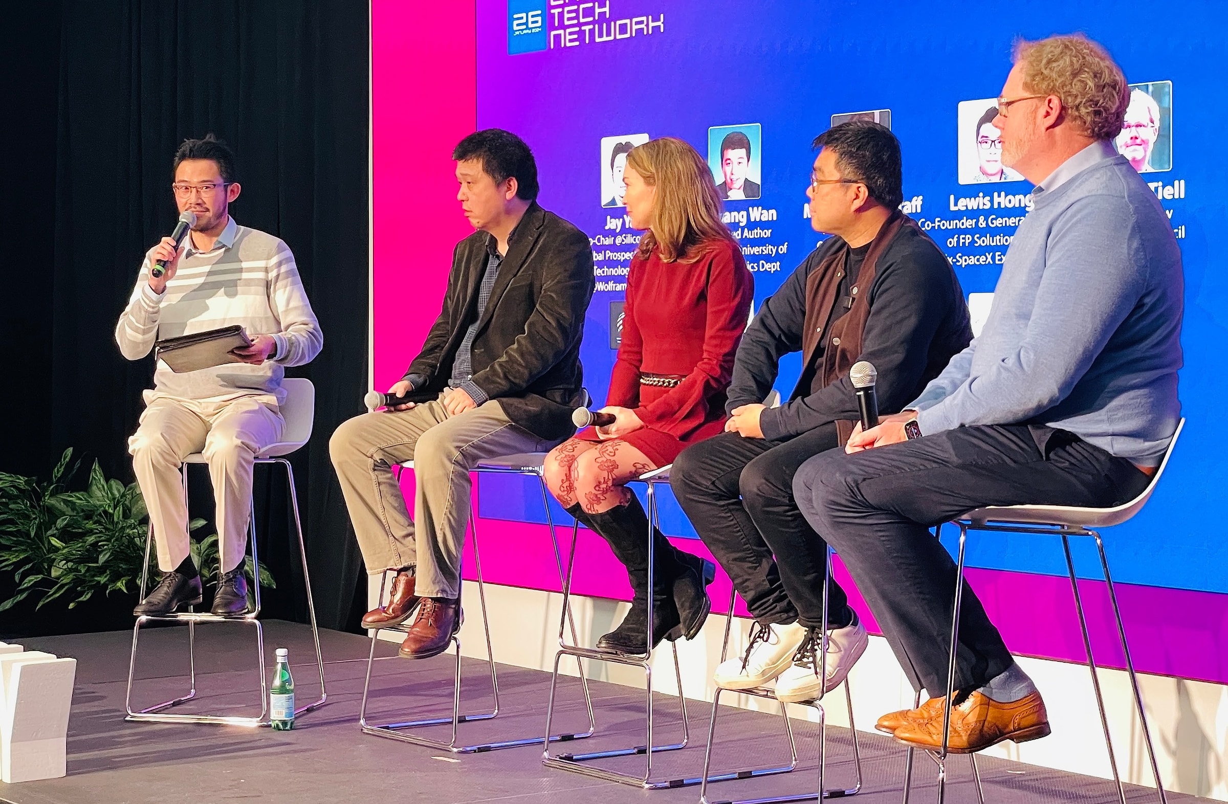 Jay moderating a panel on stage, speaking into a microphone beside four seated panelists